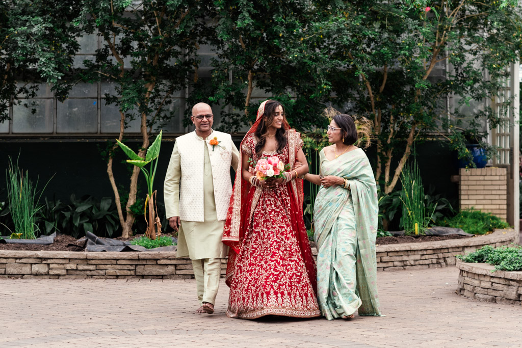 A bride in a red dress walks outside with two adults, all smiling, surrounded by greenery during a joyful Chicago Indian wedding.