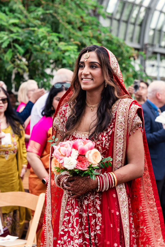 Smiling bride in red traditional attire holding a bouquet, standing outdoors with guests in the background at a vibrant Chicago Indian wedding.