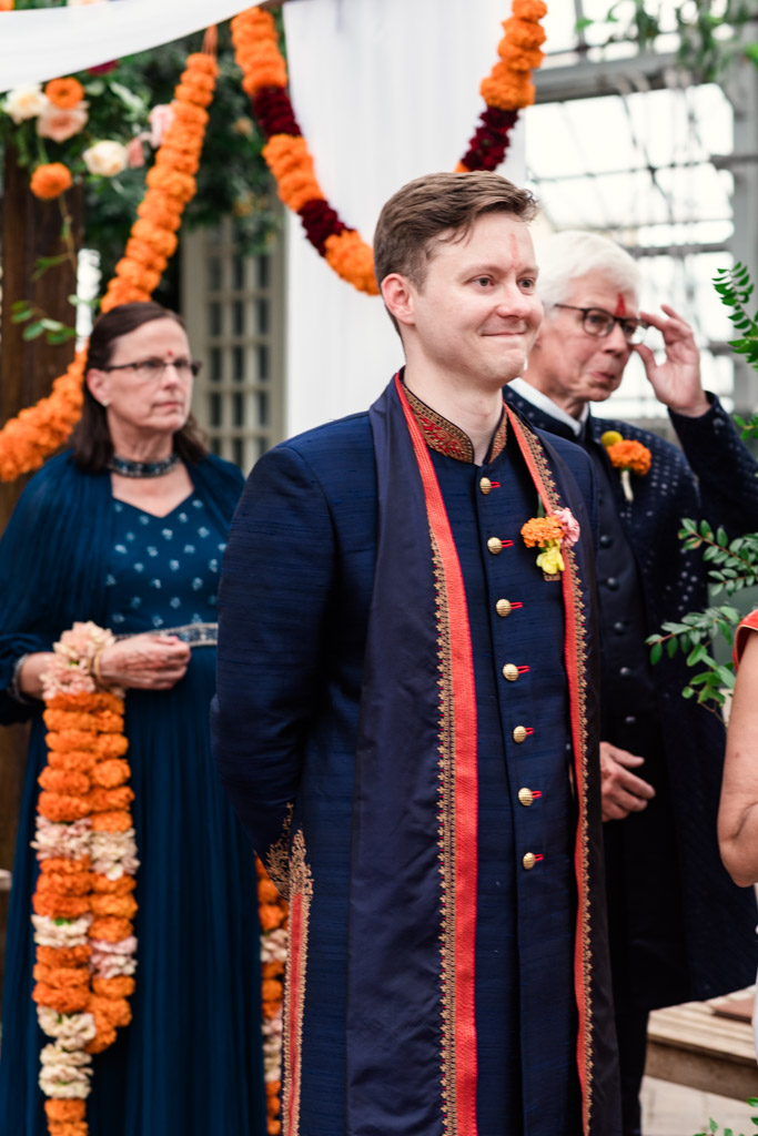 Man in traditional attire smiles at a Chicago Indian wedding ceremony, with garlands and guests in the background.