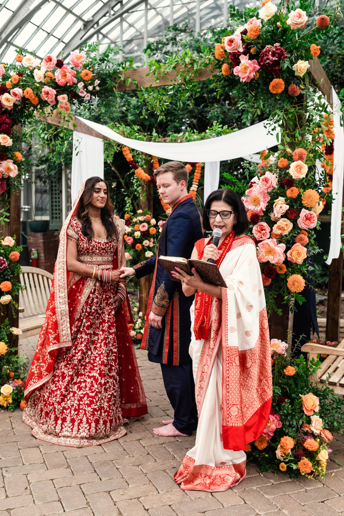 A bride and groom stand at a floral wedding altar with an officiant speaking into a microphone, capturing the elegance of a Chicago Indian wedding.