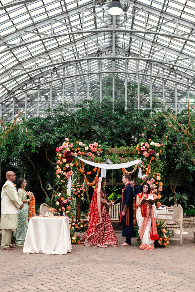 A Chicago Indian wedding ceremony in a greenhouse with a stunning floral arch, bride and groom in traditional attire, and guests nearby.