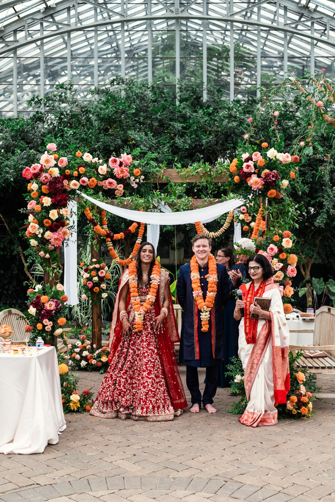 Three people in traditional attire stand under a colorful floral arch at a beautiful Chicago Indian wedding ceremony.
