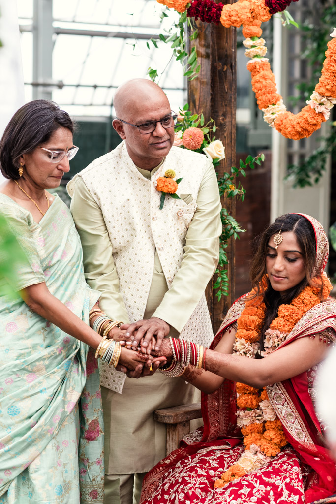 Bride in red with garlands, seated, holding hands with two adults in traditional attire at a vibrant Chicago Indian wedding, surrounded by a festive setting.