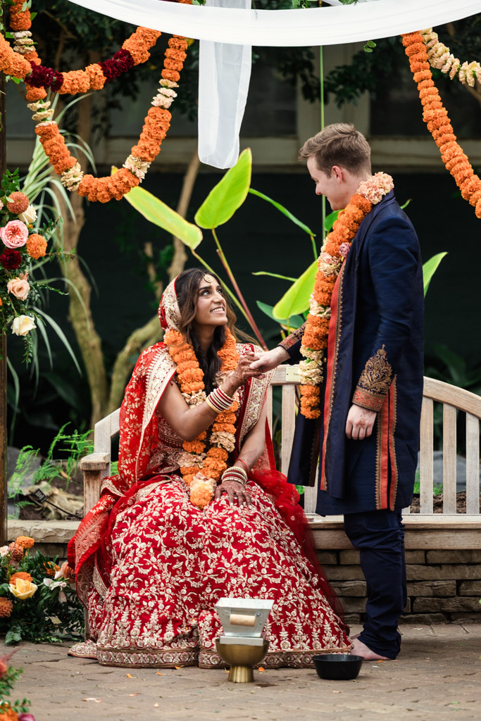 Bride in red attire sits smiling at groom in navy outfit; both wear flower garlands at their outdoor Chicago Indian wedding.