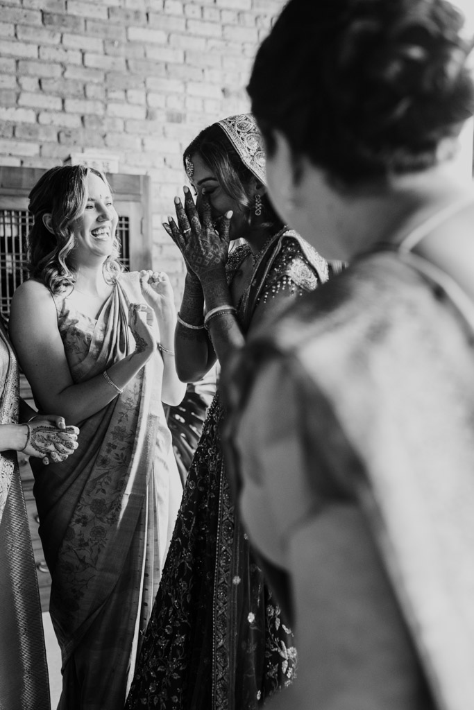Four women in sarees, one covering her face while laughing as the others smile and interact, share a candid moment at a Chicago Indian wedding.