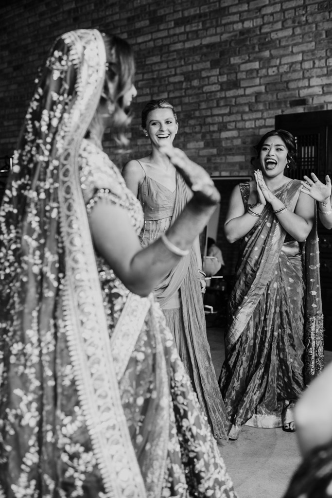 Three women in traditional attire smile and clap as they celebrate together indoors, capturing the vibrant spirit of a Chicago Indian wedding.