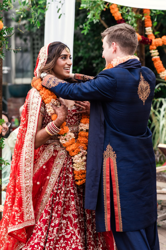 Bride in red and groom in blue exchange garlands at their Garfield Park Conservatory wedding ceremony, smiling at each other