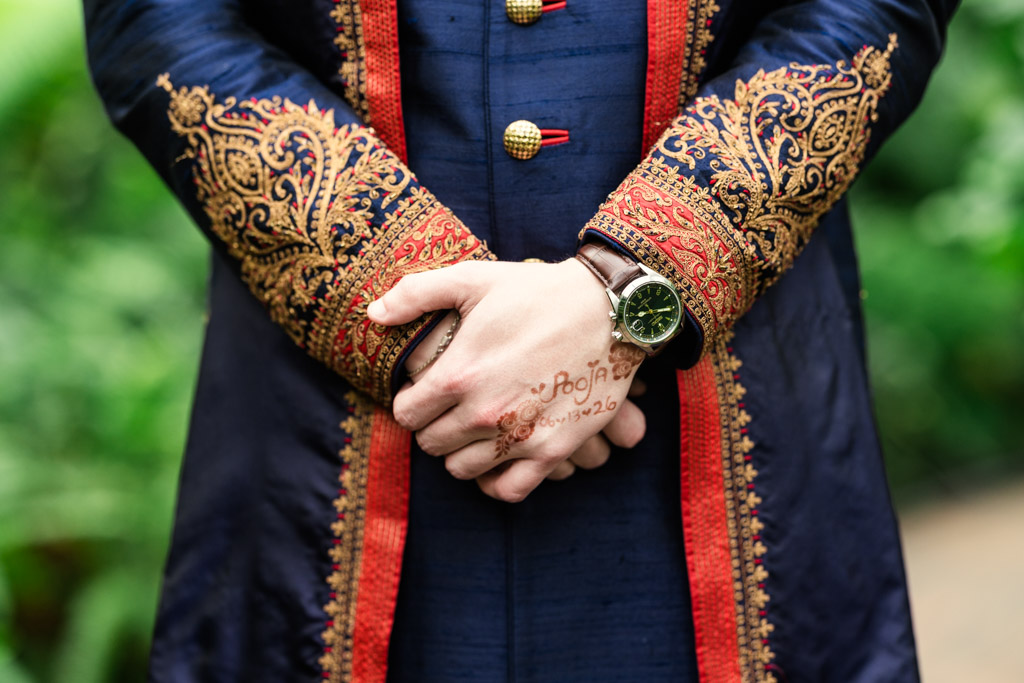 Person in an ornate navy and gold sherwani, hands clasped, wearing a watch, with intricate henna on hand, stands against lush greenery—radiating elegance at a Chicago Indian wedding.