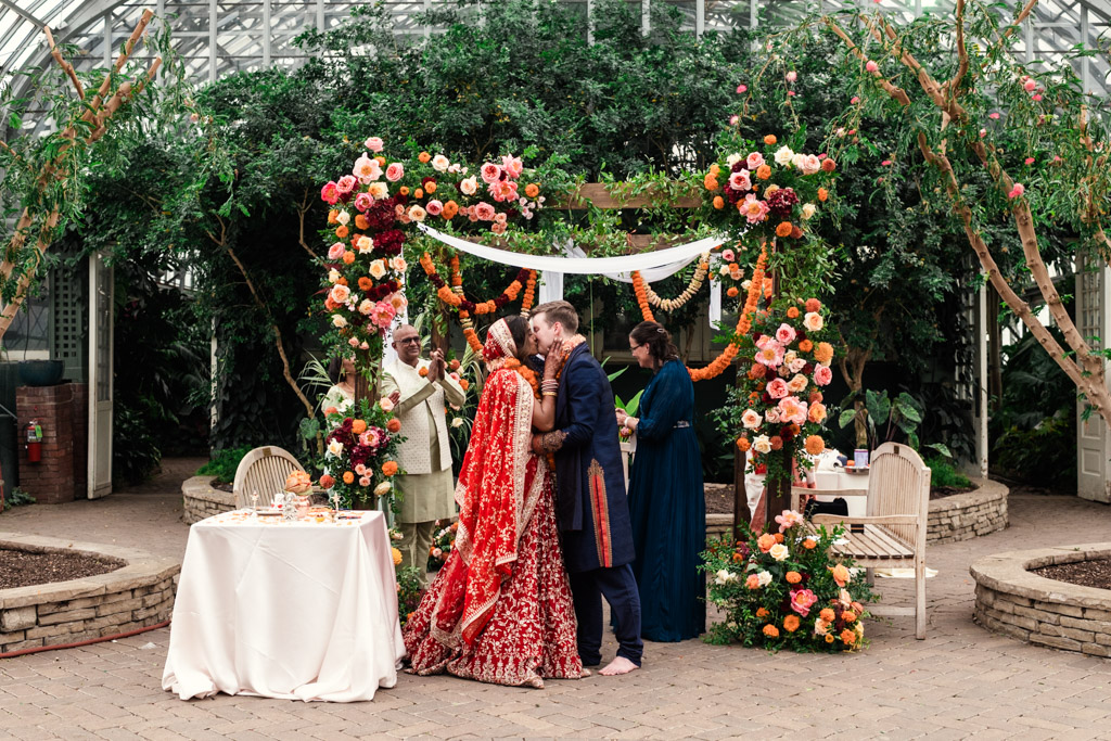 Newlywed couple kisses under a colorful floral arch during a traditional Chicago Indian wedding ceremony inside Garfield Park Conservatory