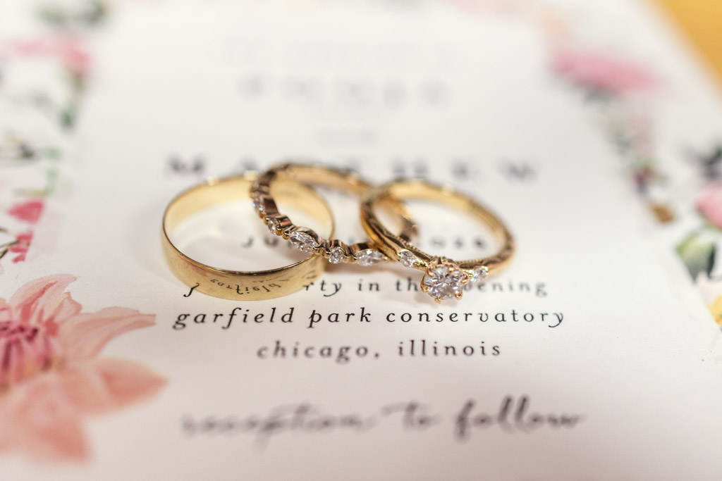Three gold rings, one with a diamond, rest on a floral wedding invitation for a Chicago Indian wedding at Garfield Park Conservatory.