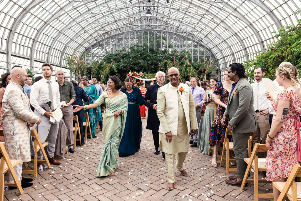 Newlyweds' parents walk down an aisle in a greenhouse surrounded by smiling guests in colorful clothing after wedding ceremony at Garfield Park Conservatory
