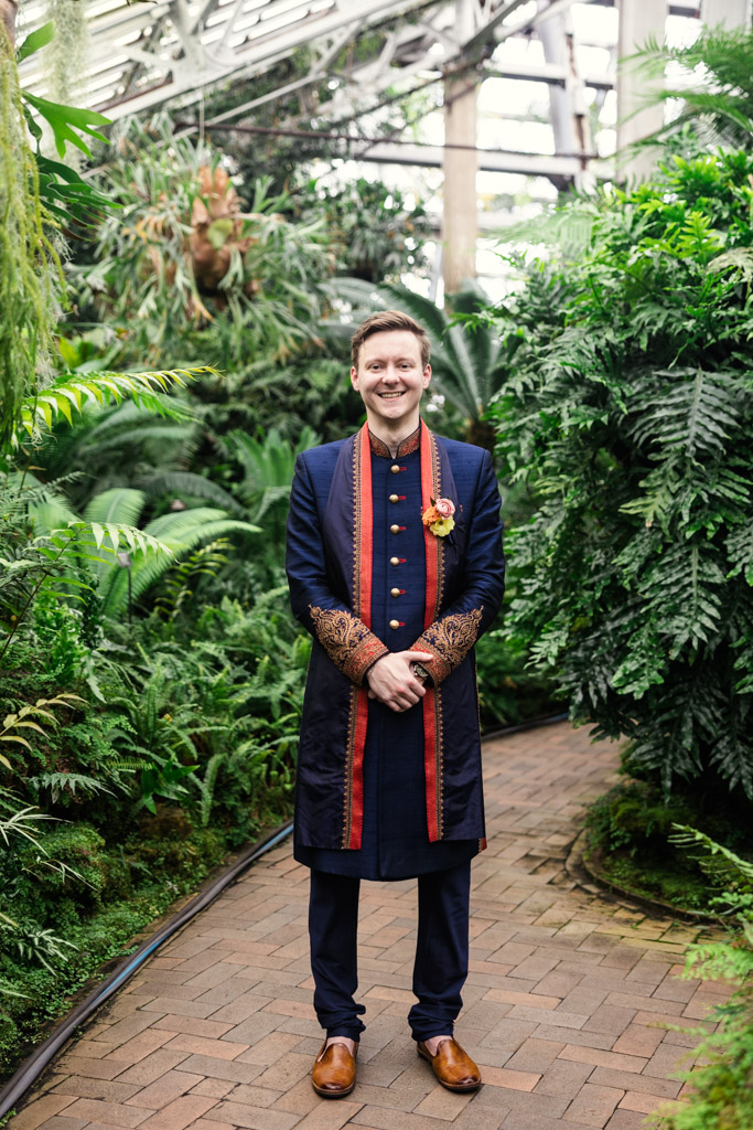 Smiling man in blue and gold sherwani stands on a brick path, surrounded by lush green plants, ready to celebrate a beautiful Chicago Indian wedding.