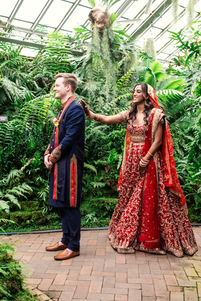 A bride in a red dress taps a groom on the shoulder in a lush, green indoor garden during their Chicago Indian wedding.