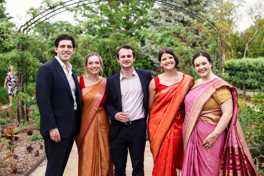 Five guests pose outdoors during a Chicago Indian wedding reception at Garfield Park Conservatory, three women in colorful saris and two men in suits, smiling at the camera