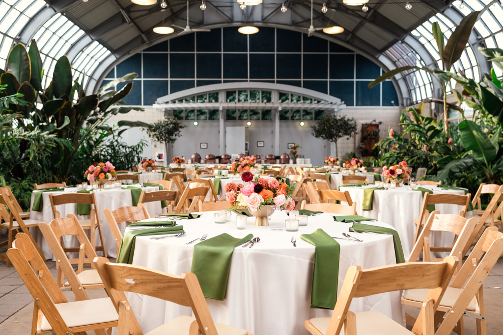 Wedding reception space at Garfield Park Conservatory with round tables with flowers and green napkins in a bright, plant-filled event space with wooden chairs