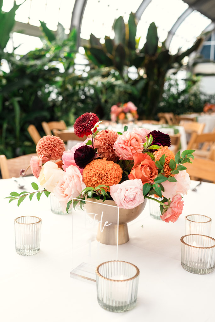Vase of pink, orange, and burgundy flowers on a table with candles in glass holders, set in a bright room for Chicago Indian wedding celebration at Garfield Park Conservatory