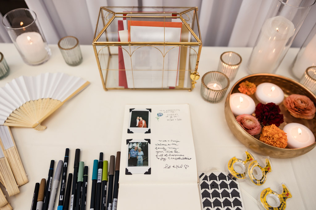 Table with pens, instant photos, a guestbook, candles, a gold box, and a bowl of flowers and floating candles for Chicago Indian wedding celebration at Garfield Park Conservatory