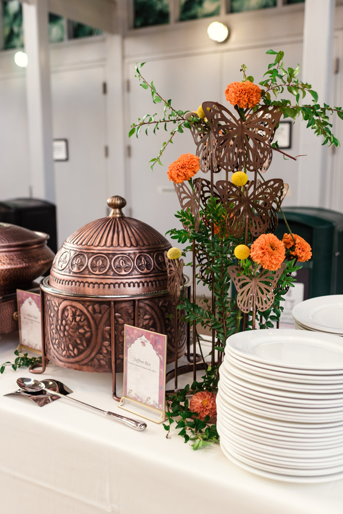 A buffet table at a Chicago Indian wedding reception features ornate copper pots, a stack of plates, and a floral decoration adorned with metal butterflies