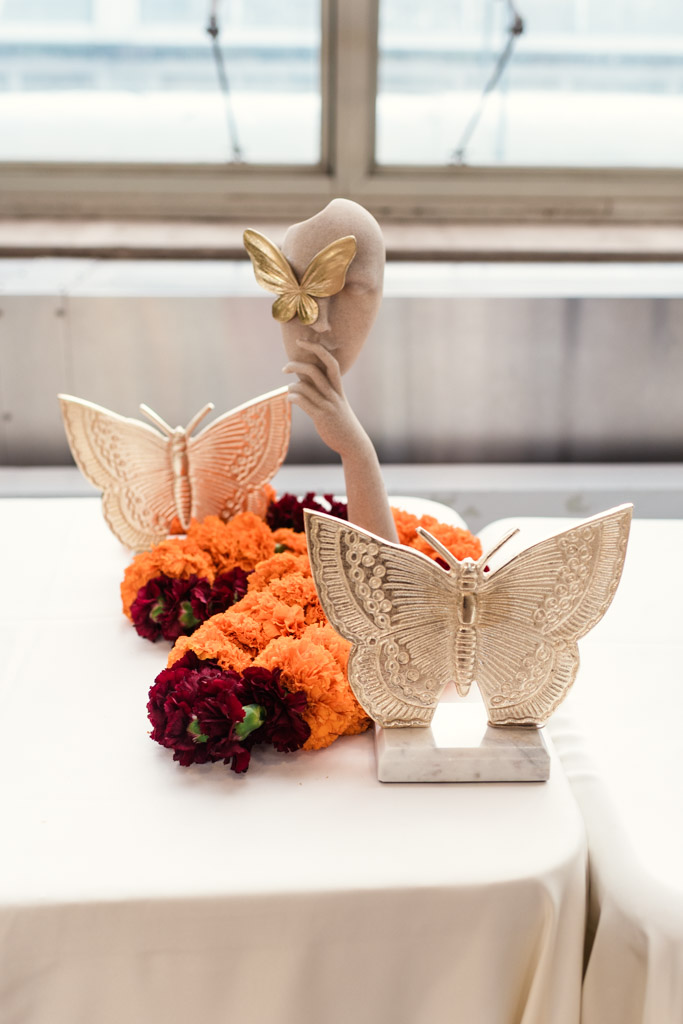 A sculpted hand holds a gold butterfly near a face, with large butterfly figures and orange flowers arranged on a table for Chicago Indian wedding at Garfield Park Conservatory
