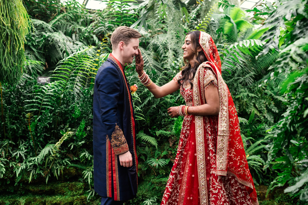A bride in red touches the groom’s face, both smiling, standing in a lush green garden at their Chicago Indian wedding.