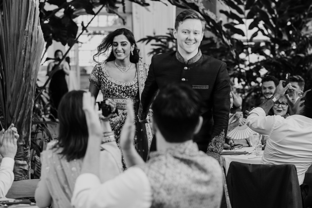 Black and white photo of newlywed couple entering their Garfield Park Conservatory wedding reception as seated guests applaud and take photos