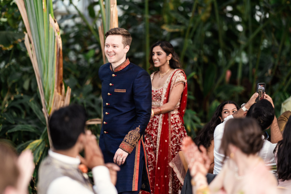 Groom in a blue sherwani and bride in a red lehenga enter their Garfield Park Conservatory wedding reception, with guests applauding