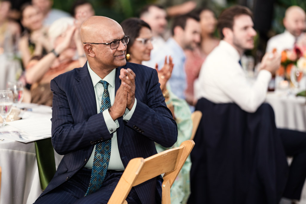 Bride's father in a suit clapping at Garfield Park Conservatory Indian wedding reception, with guests and tables in the background