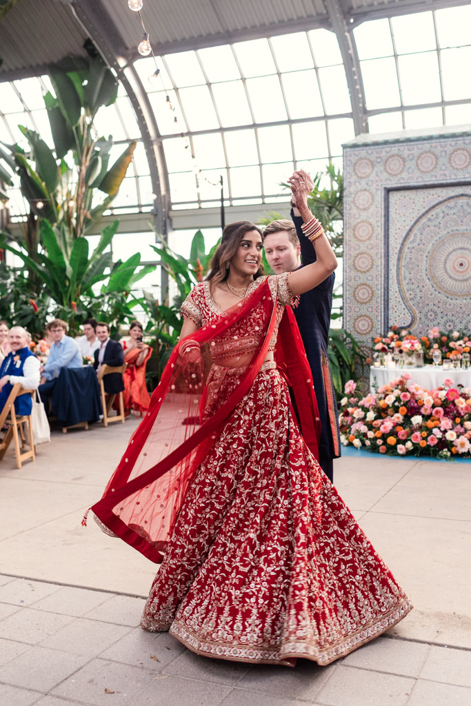 Newlywed couple dances at their wedding reception inside Garfield Park Conservatory; the bride wears a red lehenga and the venue is decorated with flowers