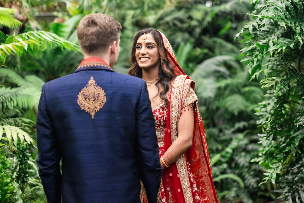 A bride in a red sari smiles at a man in a blue sherwani, surrounded by lush green plants at their Chicago Indian wedding.