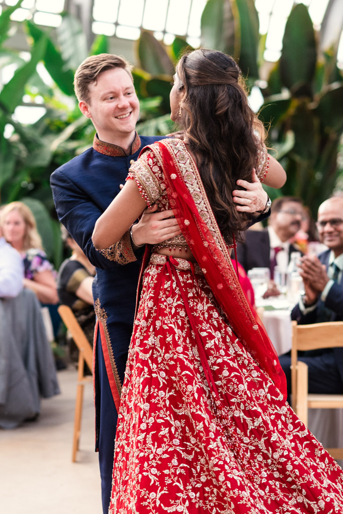 Newlywed couple dances joyfully at their Garfield Park Conservatory Indian wedding reception; bride wears a red sari, he wears blue garb, with guests watching in the background