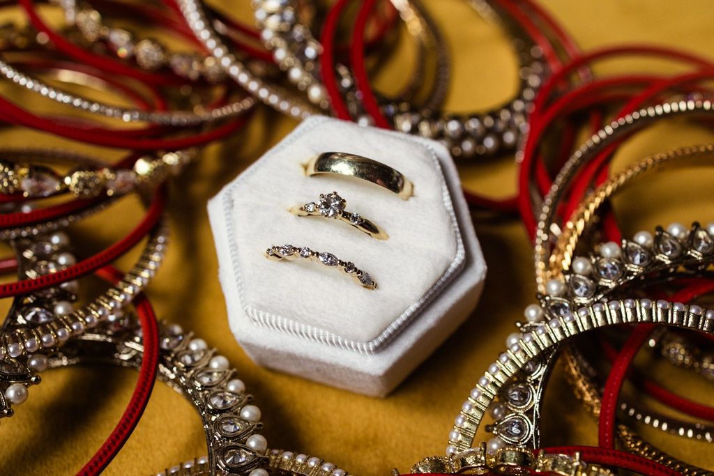 Three gold rings on a white hexagonal stand, surrounded by red and gold bangles on a yellow surface, evoke the vibrant elegance of a Chicago Indian wedding.