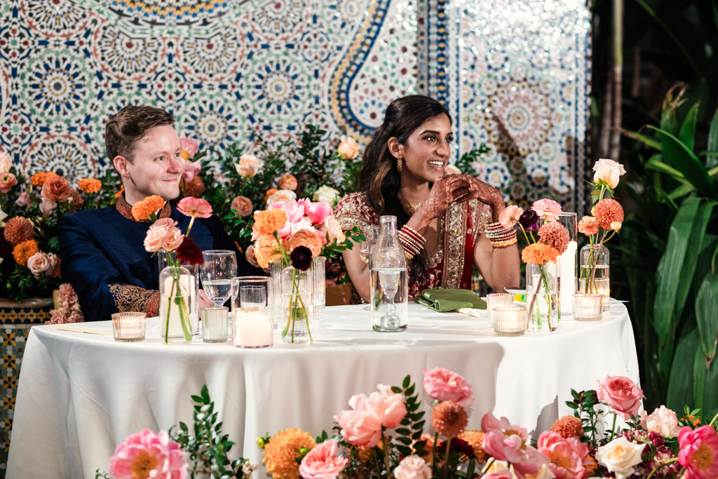 Newlywed couple sits at a flower-decorated table, smiling, with a colorful mosaic wall in the background during their Indian wedding reception at Garfield Park Conservatory