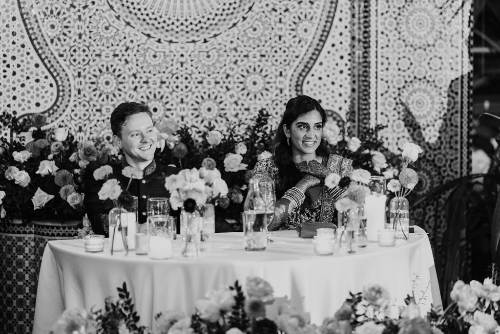 Black and white photo of happy newlyweds at a flower-adorned table with candles, in front of a tiled mosaic during their wedding reception at Garfield Park Conservatory