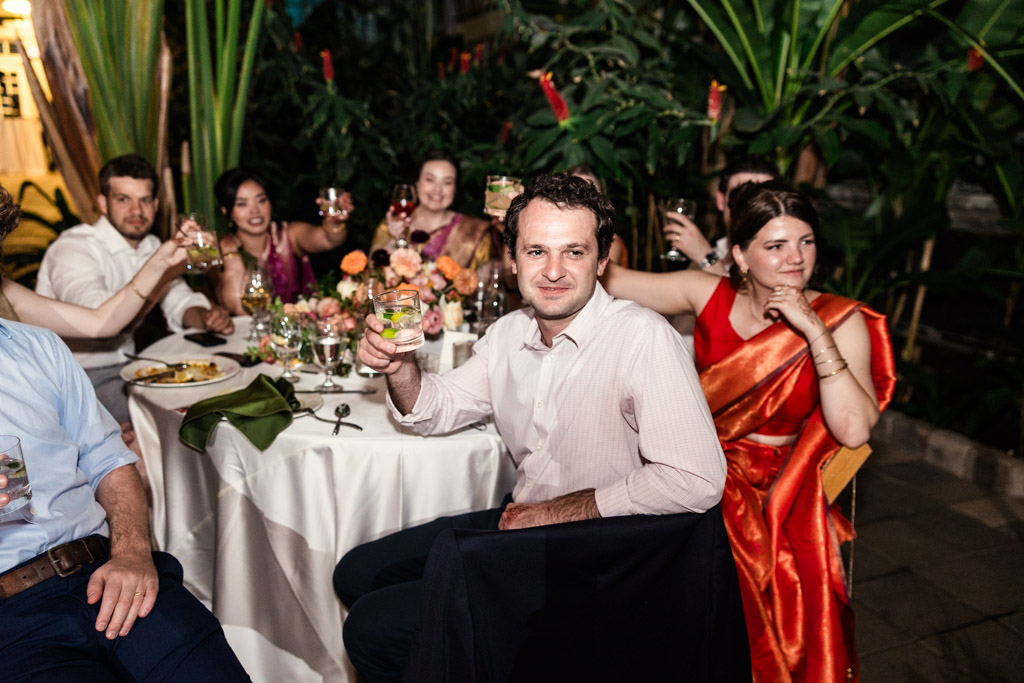 Guests in formal attire sit around a table outdoors, raising glasses in a celebratory toast, surrounded by plants during Indian wedding reception at Garfield Park Conservatory