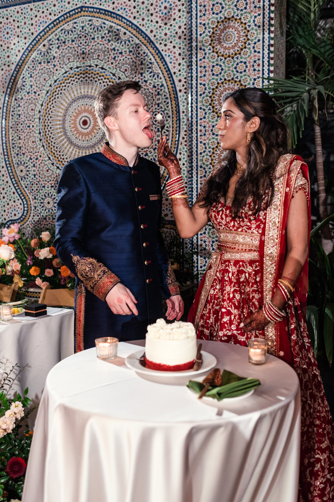Bride in red feeds cake to groom in blue amid vibrant flowers and glowing candles during their Garfield Park Conservatory wedding reception