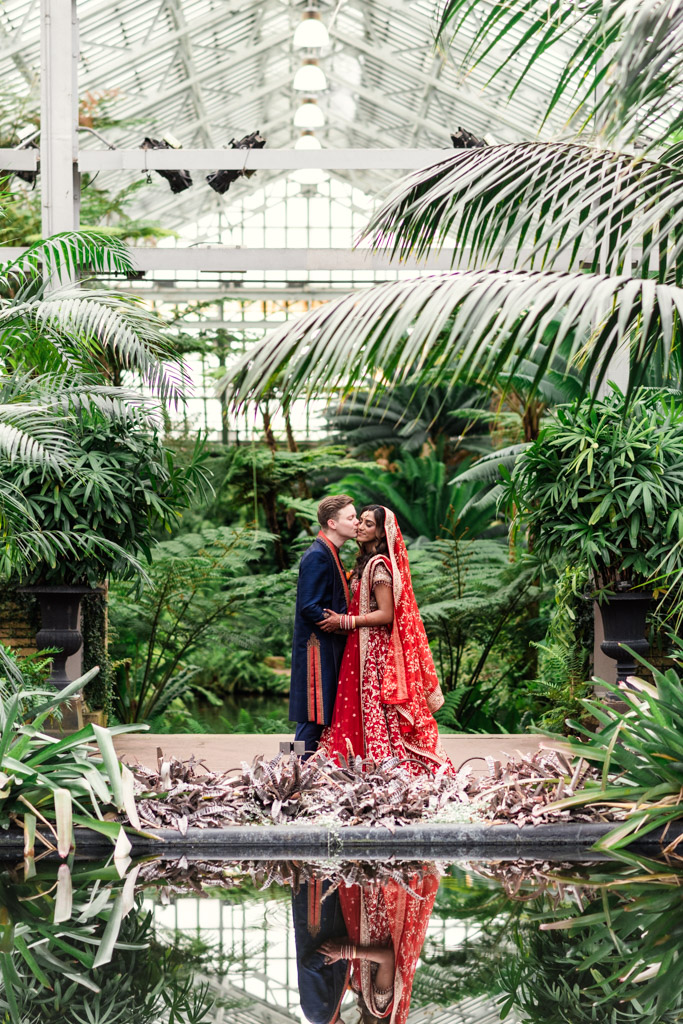 Bride and groom in traditional attire stand together in a lush, indoor botanical garden with reflection in water, capturing the elegance of a Chicago Indian wedding.