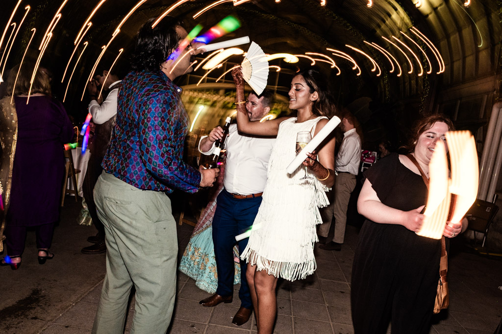 Creative photo of bride and guests dancing with fans and glow sticks at a lively Chicago Indian wedding reception, with light streaks in the background