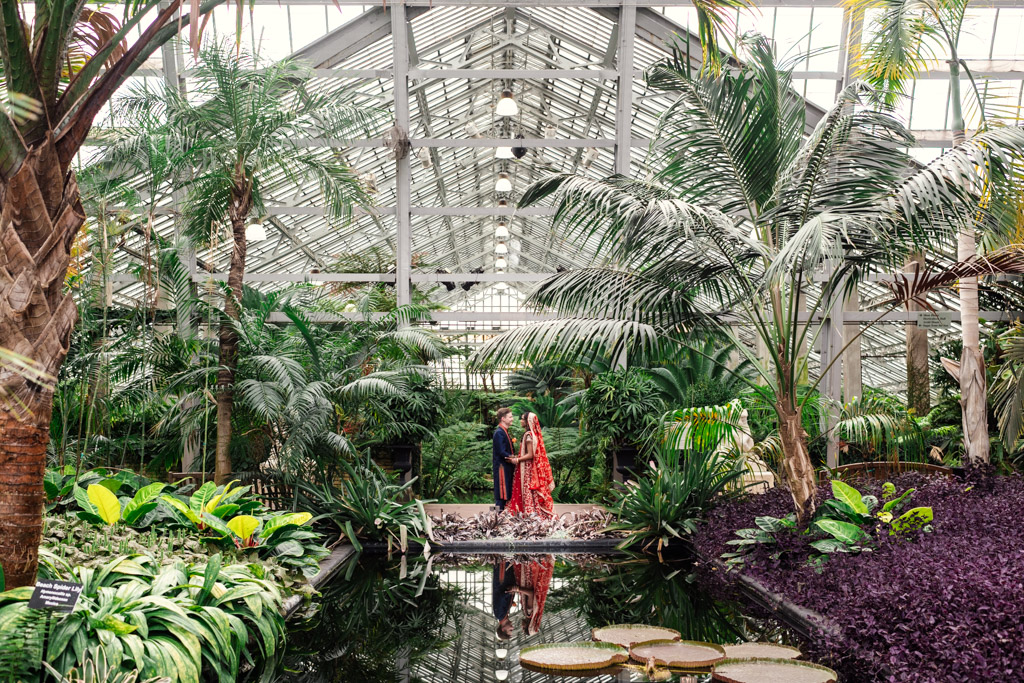 Two people stand together by a small pond in a lush indoor greenhouse filled with tropical plants, capturing a serene moment reminiscent of an elegant Chicago Indian wedding.