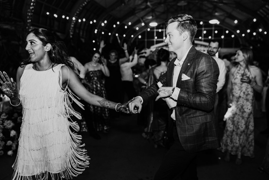 Black and white photo of newlywed couple dancing joyfully at their Chicago wedding celebration, surrounded by guests under glowing string lights at Garfield Park Conservatory