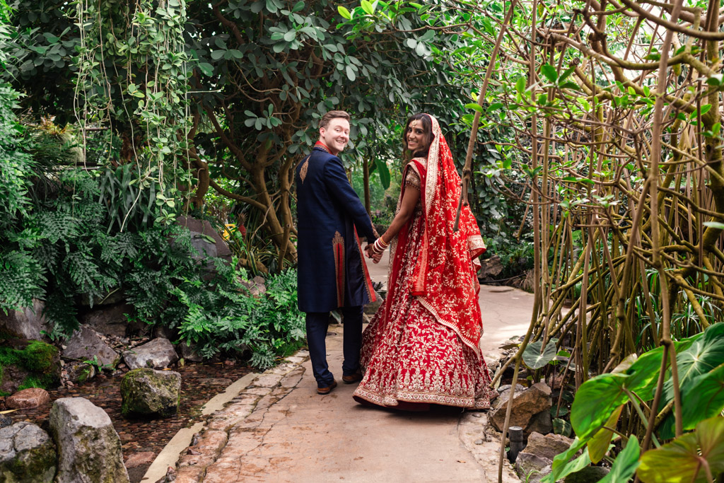 A couple in traditional attire holds hands, walking on a garden path at their Chicago Indian wedding, smiling back at the camera.