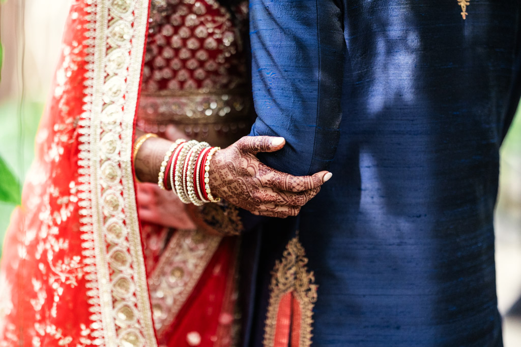 Woman in red and gold bridal attire holds arm of man in blue traditional outfit; only torsos are visible, beautifully capturing the elegance of a Chicago Indian wedding.