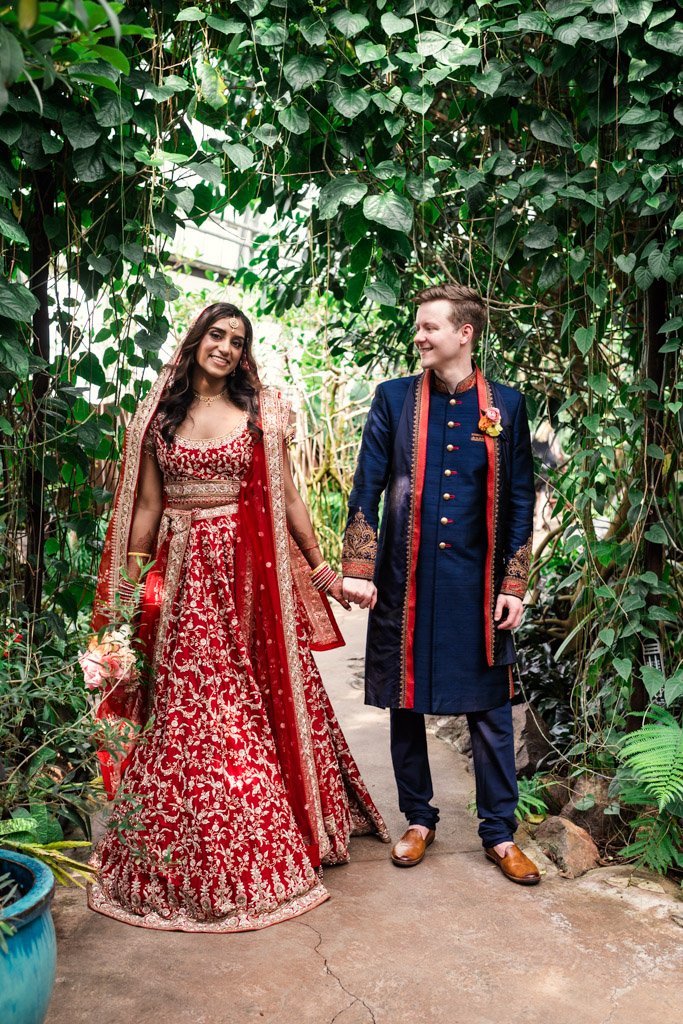 Bride in a red and gold dress and groom in a blue sherwani holding hands, standing in a lush garden at Garfield Park Conservatory