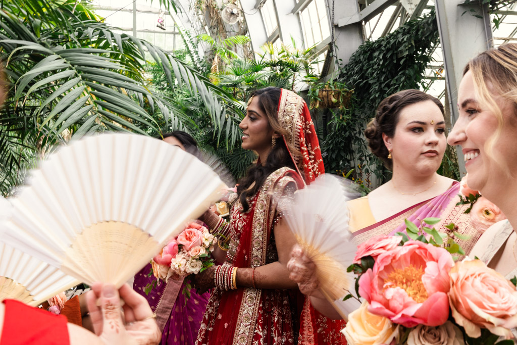 Bride in red and gold attire stands among bridesmaids with fans and flowers inside Garfield Park Conservatory