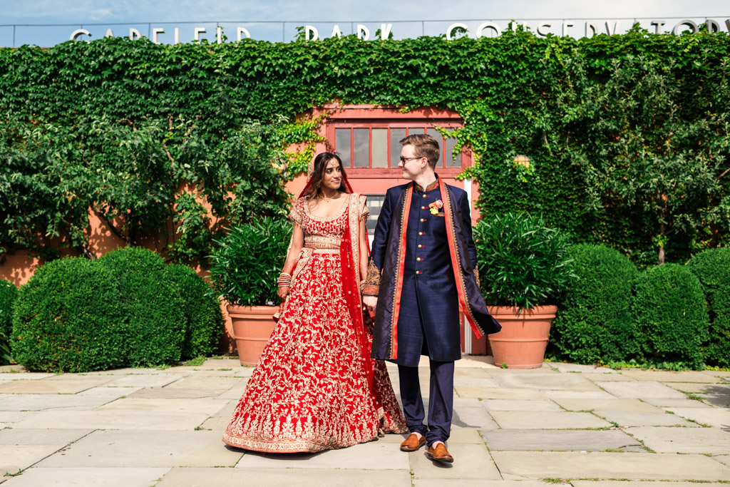 A couple in traditional Indian wedding attire holds hands and walks outdoors by a wall covered in green ivy, capturing the romantic spirit of a Chicago Indian wedding.
