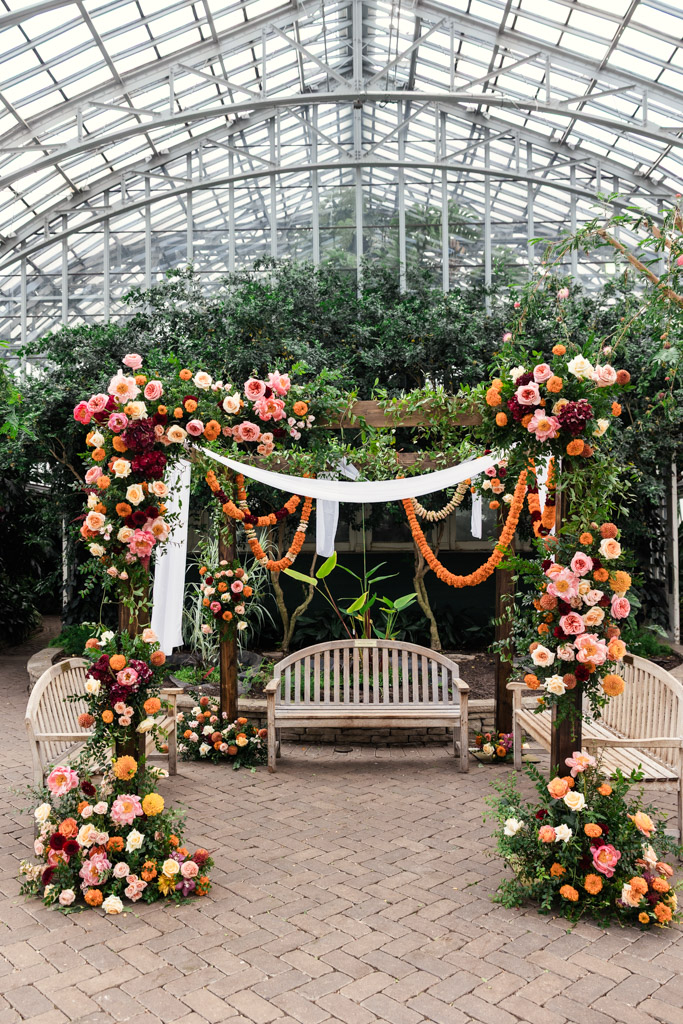 Floral arch with orange, pink, and red flowers, white fabric, and benches in a glass greenhouse setting—perfect for a romantic Chicago Indian wedding.
