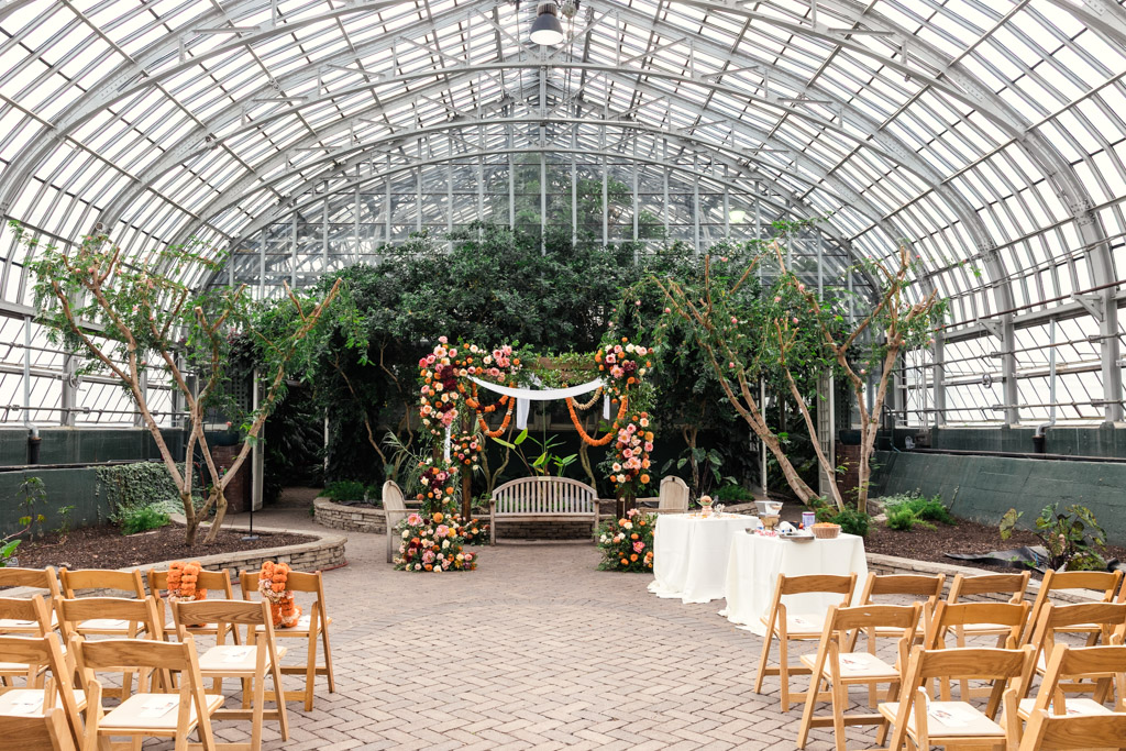 Wooden chairs face a floral arch and bench in a greenhouse set up for a Chicago Indian wedding ceremony.