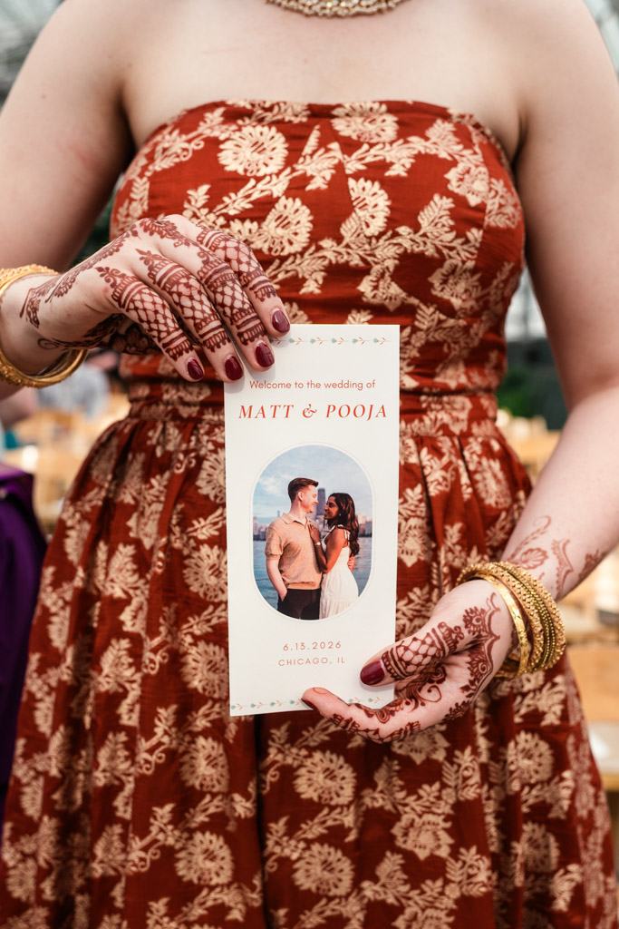 Woman in a red dress holds a wedding program with a photo and “Matt & Pooja” text, her hands adorned with henna and bangles—a scene capturing the vibrant spirit of a Chicago Indian wedding.