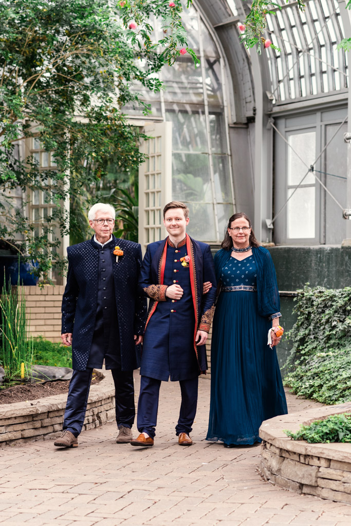 Three people in formal attire walk together in a bright, indoor garden with glass walls and lush greenery, evoking the elegance of a Chicago Indian wedding.