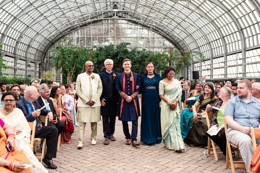Six people stand together at the front of a seated crowd in a greenhouse, dressed in formal attire, as part of an elegant Chicago Indian wedding.