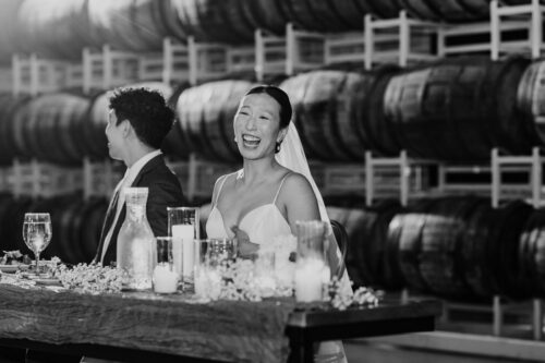 A bride laughs joyfully at a table with candles during her Barrel House wedding; barrels are stacked in the background.